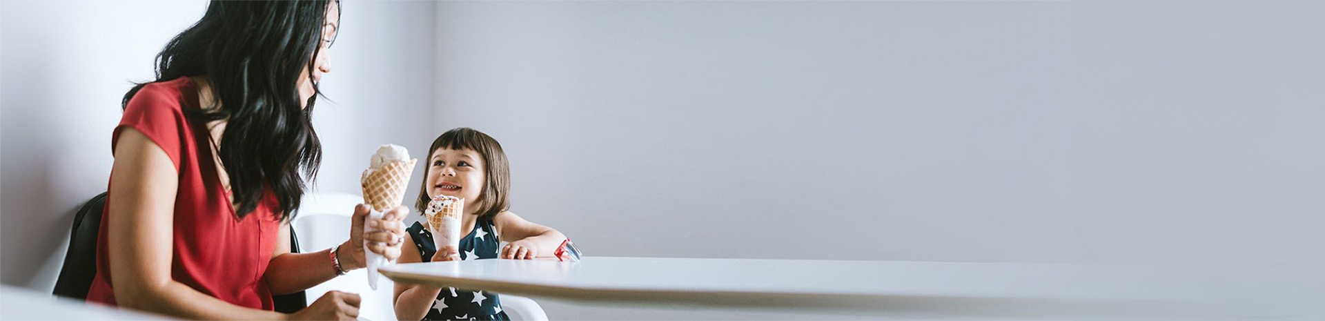 A woman and her daughter sit at a table eating ice cream cones and talking.