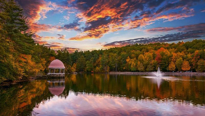 Coggshall Park in Fitchburg with a serene lake surrounded by a forest, featuring a gazebo and a fountain, reflecting autumn colors in the water.