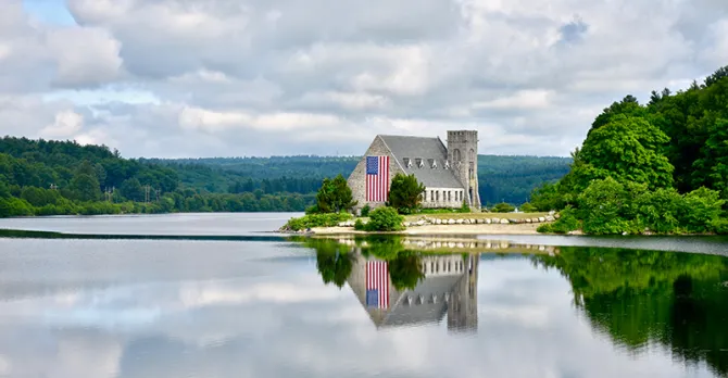 The Old Stone Church in West Boylston is shown next to a lake and cloudy skies.