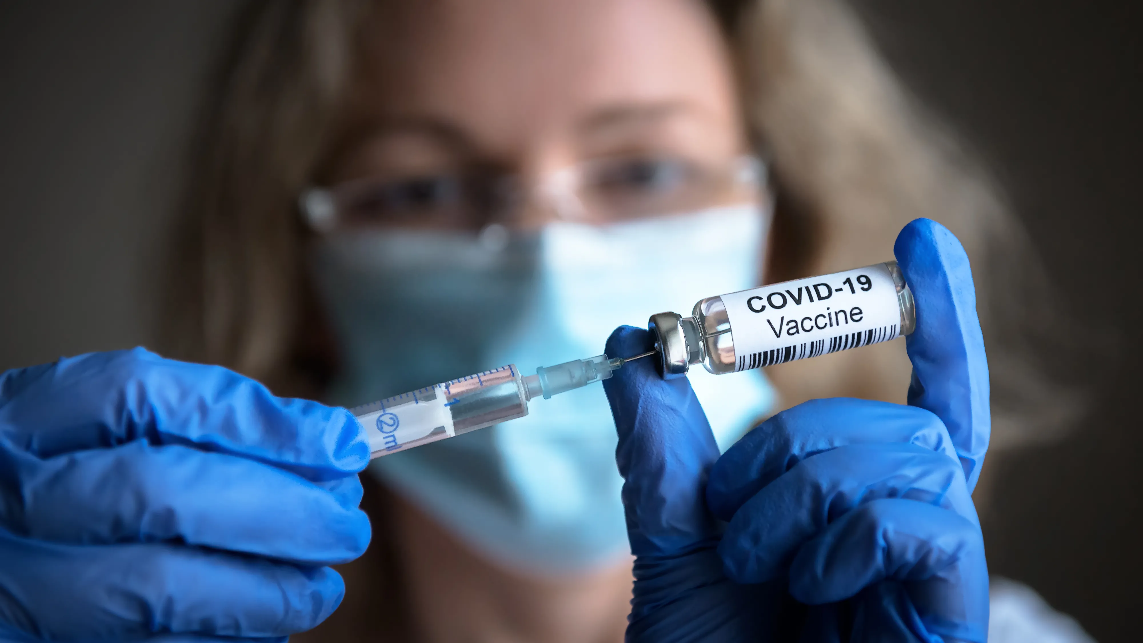 A doctor is wearing a mask and using a syringe to administer a dose of a COVID-19 vaccine.