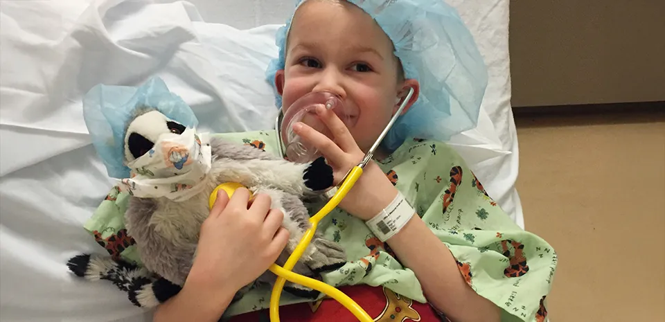 A child, holding a stuffie with a pre surgical outfit while listening to its heart with a stethoscope, has been prepped for surgery, but appears to not be scared.