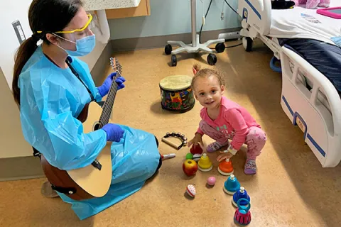 A young patient enjoys a music therapy session, playing with bells while a caregiver plays guitar.