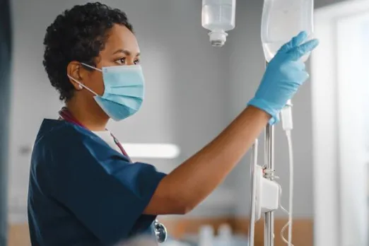 A nurse in dark teal scrubs is wearing a mask and hanging an IV bag.